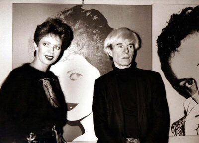 Nell'immagine si vedono, in una foto d'epoca, Regina Schrecker e Andy Warhol posare assieme davanti alla stessa opera oggi esposta all'interno della mostra "Andy Warhol - LOVE POP - Icons e Masterpieces" curata da Gianfranco Rosini, in corso a Palazzo Fulcis a Belluno.